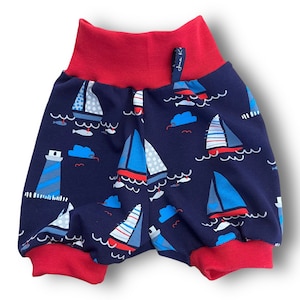 May include: Red and navy blue toddler shorts with a sailboat and lighthouse print. The shorts have a red elastic waistband and red trim around the legs.