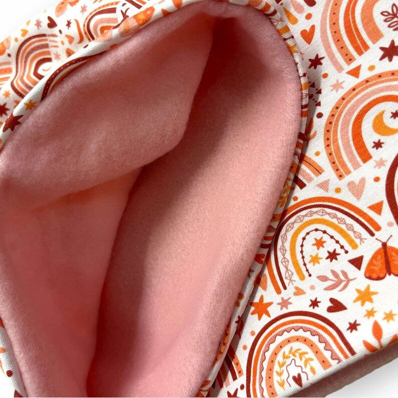 Op de afbeelding: Een roze fleece gevoerde stof met een witte achtergrond en een herhalend patroon van oranje, gele en rode regenbogen, sterren, harten en bladeren.