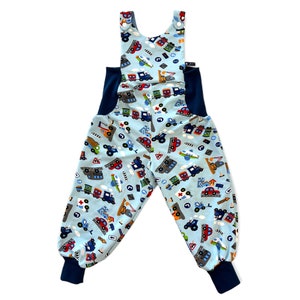 Könnte beinhalten: Blaue Latzhose mit einem Muster aus Autos, Lastwagen und Flugzeugen auf hellblauem Hintergrund. Die Latzhose hat blaue Träger und blaues Gummiband am unteren Ende der Beine.