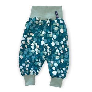 Könnte beinhalten: Babyhose mit tealfarbenem Hintergrund und einem Muster aus weißen und teal-farbenen Eukalyptusblättern. Der Bund und die Bündchen sind einfarbig salbeigrün. Ein kleines Etikett mit Text und einem Stern ist am Bund angenäht.