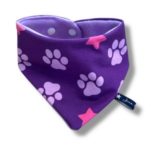 May include: A purple dog bandana with a paw print and star pattern. The bandana has a lavender interior and two white snaps. A small tag with the text "Juna" is attached to the bandana.