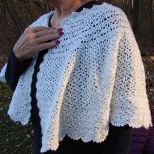 Könnte beinhalten: Ein weißer Häkelschal mit einem komplizierten durchbrochenen Muster und einem gewellten Rand. Das Kleidungsstück liegt über den Schultern und zeigt eine leichte, strukturierte Strickkonstruktion mit einem sich wiederholenden geometrischen Design.