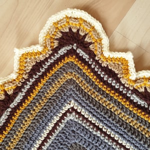 Könnte beinhalten: Ein Eckabschnitt einer quadratischen Häkeldecke mit konzentrischen Garnreihen in den Farben Dunkelgrau, Hellgrau, Weiß, Senfgelb und Dunkelbraun. Der Rand ist mit einer dekorativen, bogenförmigen Borte aus cremefarbenem Garn versehen.