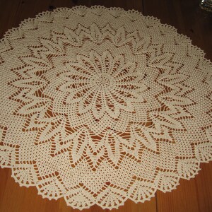 May include: A large, cream-colored crocheted doily with a delicate, intricate pattern. The doily is circular and has a scalloped edge.