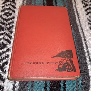 May include: A vintage hardcover book with a red cover. The title "A Judy Bolton Mystery" is printed in white. A black illustration of a tree and figures is in the lower right corner. The book is on a patterned surface.