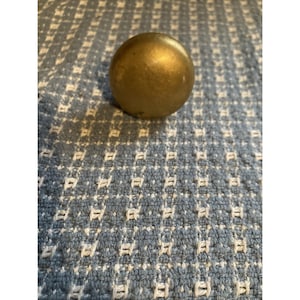 May include: A small, round, brass-colored knob. The knob is smooth and spherical, resting on a blue and white patterned fabric. The fabric has a woven texture with a repeating geometric design.