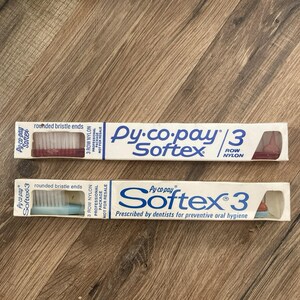 May include: Two vintage Py-co-pay Softex 3 row nylon toothbrushes in their original packaging. One has red bristles, the other has blue bristles. The packaging is white with blue text and a small window showing the brush.