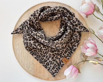 May include: A leopard print scarf in shades of brown and black, arranged on a wooden tray. The scarf is triangular and has a tag attached. Pink and white magnolia flowers are in the background.
