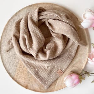 May include: A beige muslin baby bandana with white polka dots. The bandana is folded and displayed on a wooden surface. The bandana has a small brown tag with the text "Made with Love" on it.