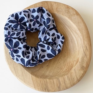 Può includere: Un elastico per capelli azzurro con un motivo floreale blu scuro e piccoli accenti rosa. L'elastico è appoggiato in una ciotola rotonda di legno con finitura marrone chiaro. La ciotola è su una superficie bianca.