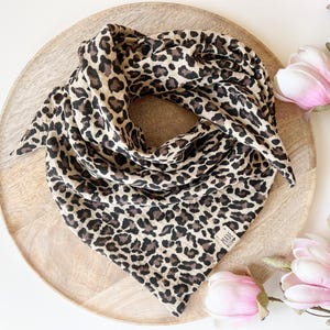 May include: A leopard print scarf in tan, brown, and black, displayed on a wooden tray. The scarf is folded into a triangle shape. Pink and white flowers are in the background. The scarf has a small tag that says "MADE WITH LOVE".