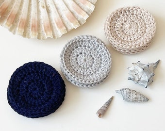 May include: Three crocheted coasters in different colors: navy blue, light gray, and beige. The coasters are round and have a textured surface. There are also three seashells on the white surface.