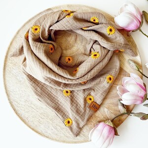 Könnte beinhalten: Ein beigefarbener Musselin-Schal mit gestickten gelben Sonnenblumen. Der Schal liegt auf einem runden Holztablett, im Hintergrund rosa und weiße Magnolienblüten. Ein kleines braunes Etikett ist angebracht.