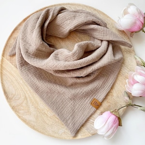 Può includere: Sciarpa in mussola beige con motivo a pois, appoggiata su un vassoio di legno. La sciarpa è piegata a forma di triangolo. Una piccola etichetta marrone è attaccata alla sciarpa. Fiori di magnolia rosa e bianchi sullo sfondo.