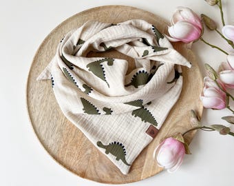 May include: A beige muslin baby bib with a green dinosaur pattern. The bib is triangular and folded on a wooden tray, with pink and white flowers in the background. The bib has a small brown leather tag.