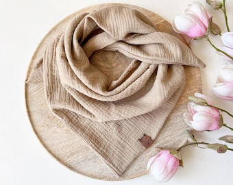 May include: A beige muslin scarf is arranged on a wooden tray, with a small fox-shaped tag. Pink and white magnolia flowers are placed around the scarf. The scarf has a textured, crinkled appearance.