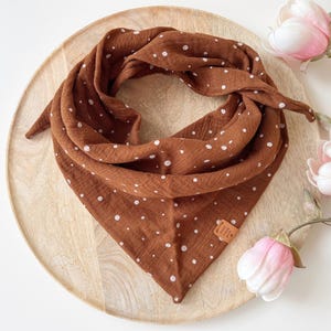 Op de afbeelding: Een bruine sjaal met witte stippen, in een driehoekige vorm, ligt op een houten dienblad. De sjaal heeft witte stippen en een klein bruin leren label. Roze en witte bloemen zijn op de achtergrond.