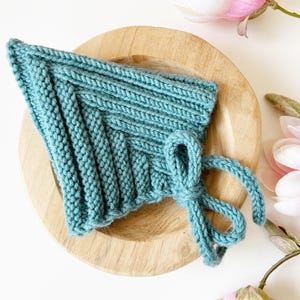 Puede incluir: Un gorro de bebé tejido en color turquesa con cierre de lazo, sobre un plato de madera clara. El gorro presenta un diseño de punto acanalado y está rodeado de flores y capullos de magnolia rosa.