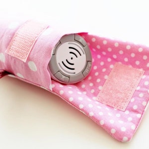 May include: A pink polka dot fabric pouch with a velcro closure. The pouch is designed to hold a small, round, gray electronic device with buttons and a white symbol.