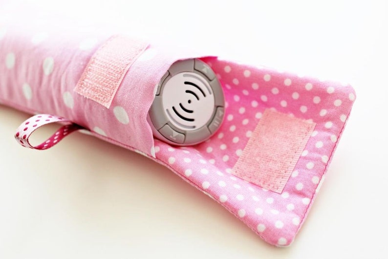 May include: A pink polka dot fabric pouch with a velcro closure. The pouch is designed to hold a small, round, gray electronic device with buttons and a white symbol.