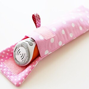 May include: A pink polka dot fabric pouch with a white velcro closure and a pink and white ribbon loop. The pouch is holding a white and orange electronic device with buttons.