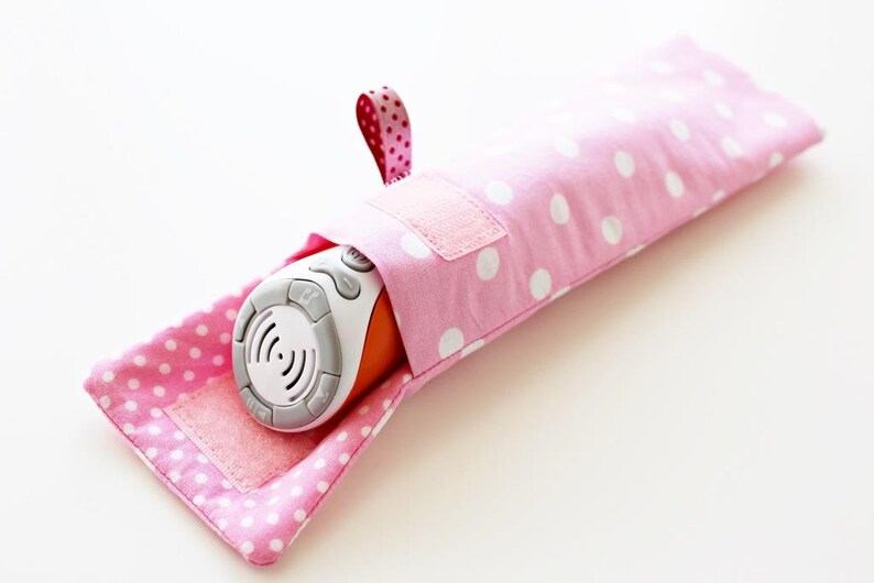 May include: A pink polka dot fabric pouch with a white velcro closure and a pink and white ribbon loop. The pouch is holding a white and orange electronic device with buttons.
