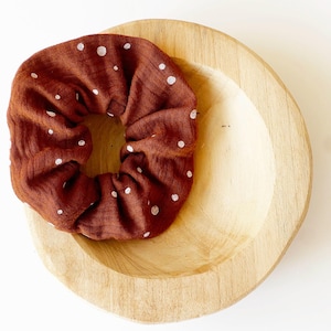 Può includere: Un elastico per capelli in tessuto color ruggine con un motivo a piccoli pois bianchi. L'elastico ha un aspetto arricciato e testurizzato ed è appoggiato all'interno di una ciotola in legno chiaro e liscio, di forma circolare e concava.