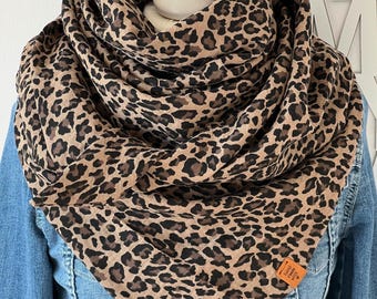 May include: A brown and black leopard print infinity scarf. The scarf is wrapped around a white mannequin head and worn over a denim jacket. A small orange tag is sewn on the scarf.
