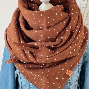 Puede incluir: Una bufanda de color óxido con lunares blancos está drapeada alrededor de una cabeza de maniquí y una chaqueta vaquera. La bufanda está hecha de una tela suave y texturizada. Una pequeña etiqueta rectangular está adherida a la bufanda.
