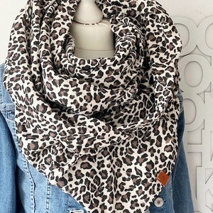 Puede incluir: Bufanda infinita con estampado de leopardo en tonos marrón, negro y blanco, drapeada sobre una chaqueta vaquera. La bufanda está hecha de tela suave y presenta un patrón de estampado animal repetido. Una pequeña etiqueta de cuero marrón está cosida en la bufanda.