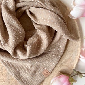 May include: A brown muslin baby swaddle with white polka dots. The swaddle is folded and displayed on a wooden surface with a small brown leather tag that says "Made with Love".