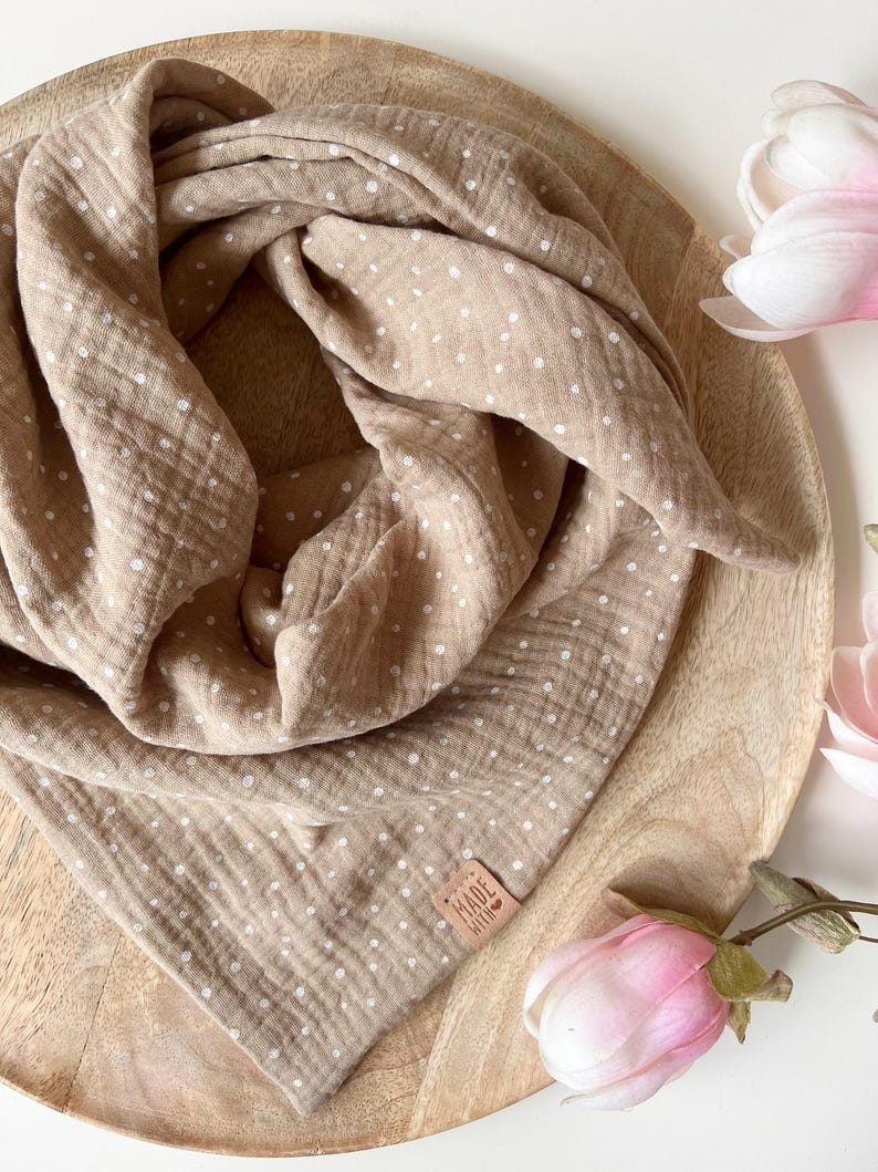 May include: A brown muslin baby swaddle with white polka dots. The swaddle is folded and displayed on a wooden surface with a small brown leather tag that says "Made with Love".