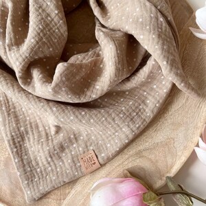 May include: A brown muslin baby swaddle with white polka dots. The swaddle is folded and displayed on a wooden surface. A small leather tag with the text "MADE WITH LOVE" is attached to the swaddle.