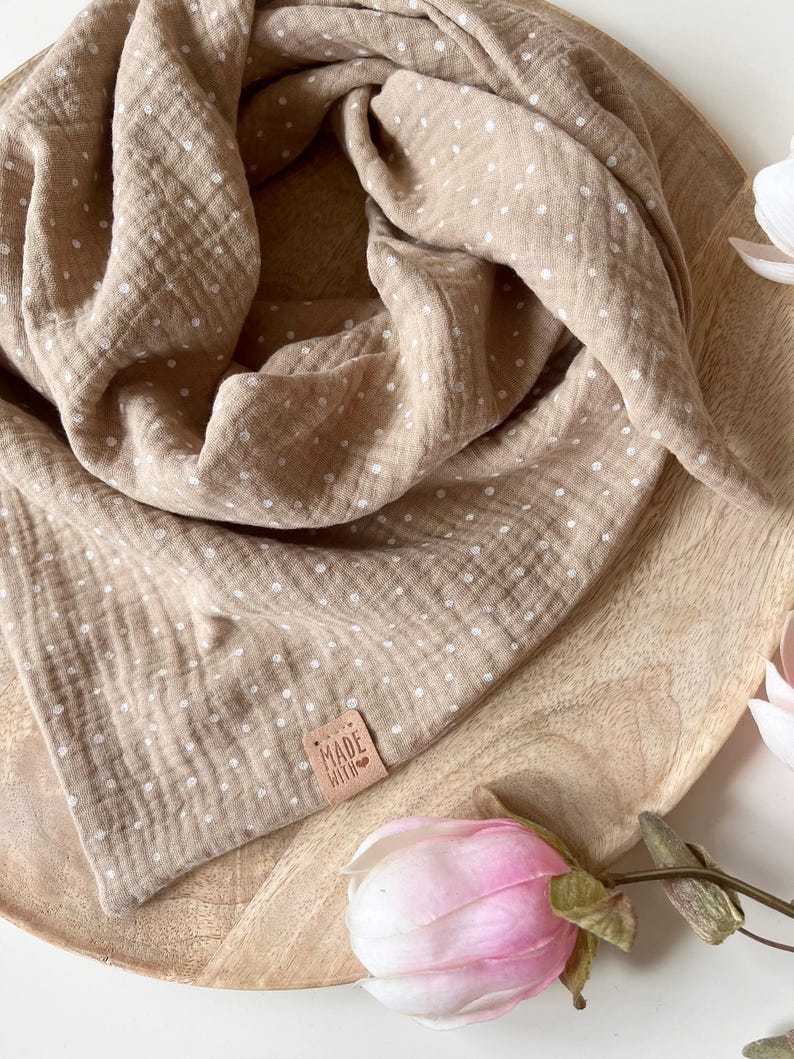 May include: A brown muslin baby swaddle with white polka dots. The swaddle is folded and displayed on a wooden surface. A small leather tag with the text "MADE WITH LOVE" is attached to the swaddle.