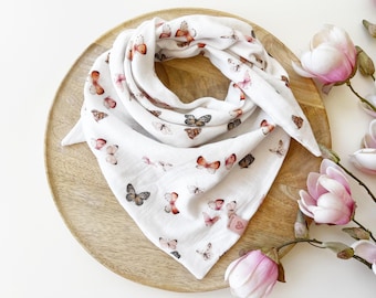 May include: A white triangular scarf featuring a repeating pattern of butterflies in shades of orange, brown, and black. The fabric has a soft, crinkled texture and includes a small, square, pink fabric tag with a heart icon on the bottom corner.