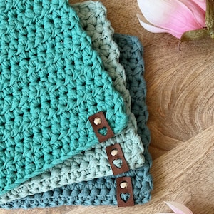 Op de afbeelding: Vier gehaakte onderzetters in teal, blauw en groen met bruine leren accenten in de vorm van een hart. De onderzetters zijn gestapeld op een houten oppervlak.