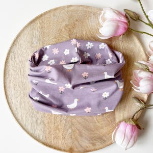 May include: A lilac neck warmer featuring a pattern of white ducks and small white and pink flowers. The neck warmer is folded on a wooden tray, with pink and white flowers in the background.