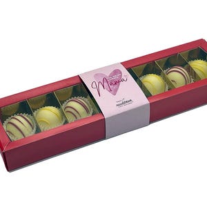 Op de afbeelding: Een rode rechthoekige doos met zes witte chocoladetruffels. Elke truffel zit in een papieren cup. Sommige truffels hebben rode strepen. De doos heeft een roze band met het woord "Mama" en een hart.