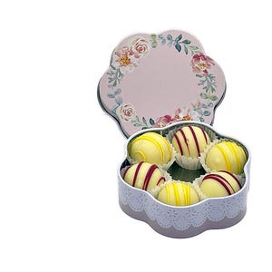 Peut inclure: Une boîte cadeau en forme de fleur avec un couvercle floral, ouverte pour révéler six truffes au chocolat blanc rondes. Les truffes ont des rayures jaunes et rouges. La boîte est rose clair avec une bordure en dentelle blanche.