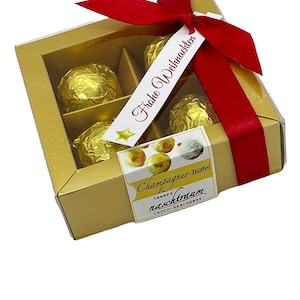 Peut inclure: Une boîte cadeau dorée avec une fenêtre transparente, contenant quatre truffes au chocolat enveloppées d'or. La boîte est attachée avec un ruban rouge et une étiquette qui dit "Frohe Weihnachten". Une étiquette sur la boîte indique "Champagner Trüffel".