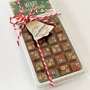 Può includere: Un calendario dell'Avvento al cioccolato in una confezione di plastica trasparente. Contiene 24 quadratini numerati con decorazioni festive. La confezione è legata con spago rosso e bianco e include un'etichetta e una carta con "Merry Christmas".