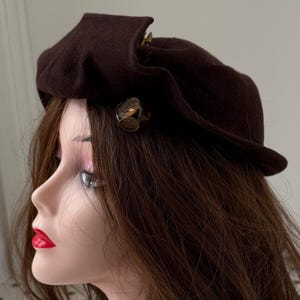 May include: A dark brown beret with a small brim and a decorative gold-colored clasp. The beret is displayed on a mannequin head with long brown hair and red lipstick. The hat is a classic accessory.