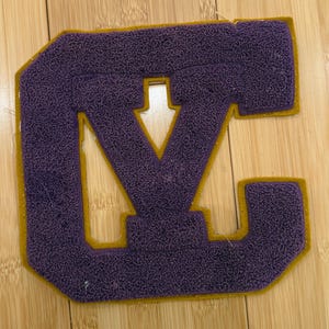 May include: A purple felt letter patch with a gold border. The patch features the letters "VC" in a varsity style. The patch is likely for a jacket or other apparel item.