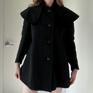 May include: A black coat with a unique cape-like collar and three large buttons down the front. The coat has long sleeves and a flared shape, creating a vintage aesthetic. The coat is made of a textured fabric.