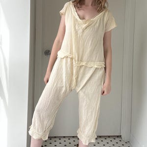 Könnte beinhalten: Cremefarbenes zweiteiliges Leinen-Outfit. Das Oberteil hat kurze Ärmel und einen gerüschten Ausschnitt. Die Hose ist cropped, mit gerüschtem Saum. Das Outfit wird mit schwarzen Stiefeln und bunten Socken getragen. Der Hintergrund ist ein weißer Flur mit einer Tür.