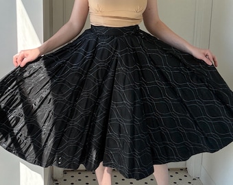 May include: A beige crop top with short sleeves and a scoop neckline paired with a black skirt. The skirt has a full, flared silhouette and features a white, wavy pattern. The outfit is suitable for a vintage or retro aesthetic.