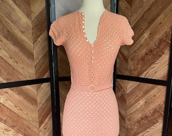 May include: A peach-colored crochet dress with a V-neckline and short sleeves. The dress has a belt and button closures.