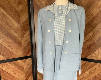 Puede incluir: Un traje de tres piezas azul claro con una chaqueta de doble botonadura, una blusa de cuadros vichy y una falda. El traje está hecho de tela de lino.