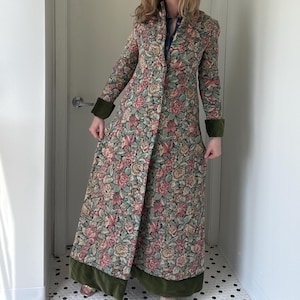 Op de afbeelding: Lange bloemenjas met knoopsluiting, met een patroon van roze, groene en beige bloemen. De jas heeft donkergroene fluwelen afwerking op de manchetten en zoom. De jas wordt gedragen over een spijkerblouse.