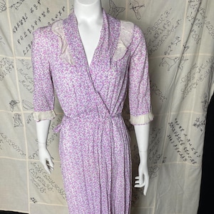 Könnte beinhalten: Ein Wickelkleid mit floralem Muster in Lila und Weiß mit V-Ausschnitt und 3/4-Ärmeln. Das Kleid hat einen weißen Spitzenbesatz an den Ärmeln und am Ausschnitt.